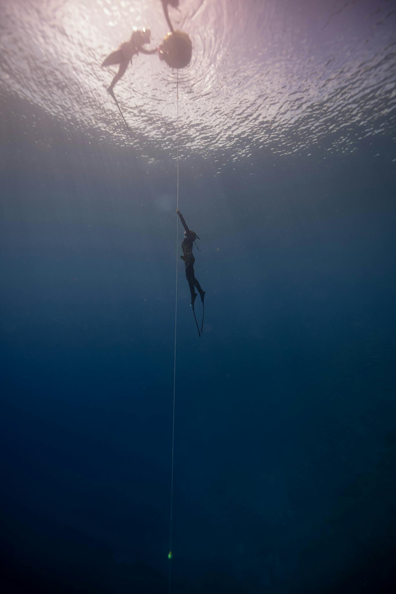 Side view of anonymous diver in wetsuit and flippers swimming up rope under transparent crystal blue seawater close to surface