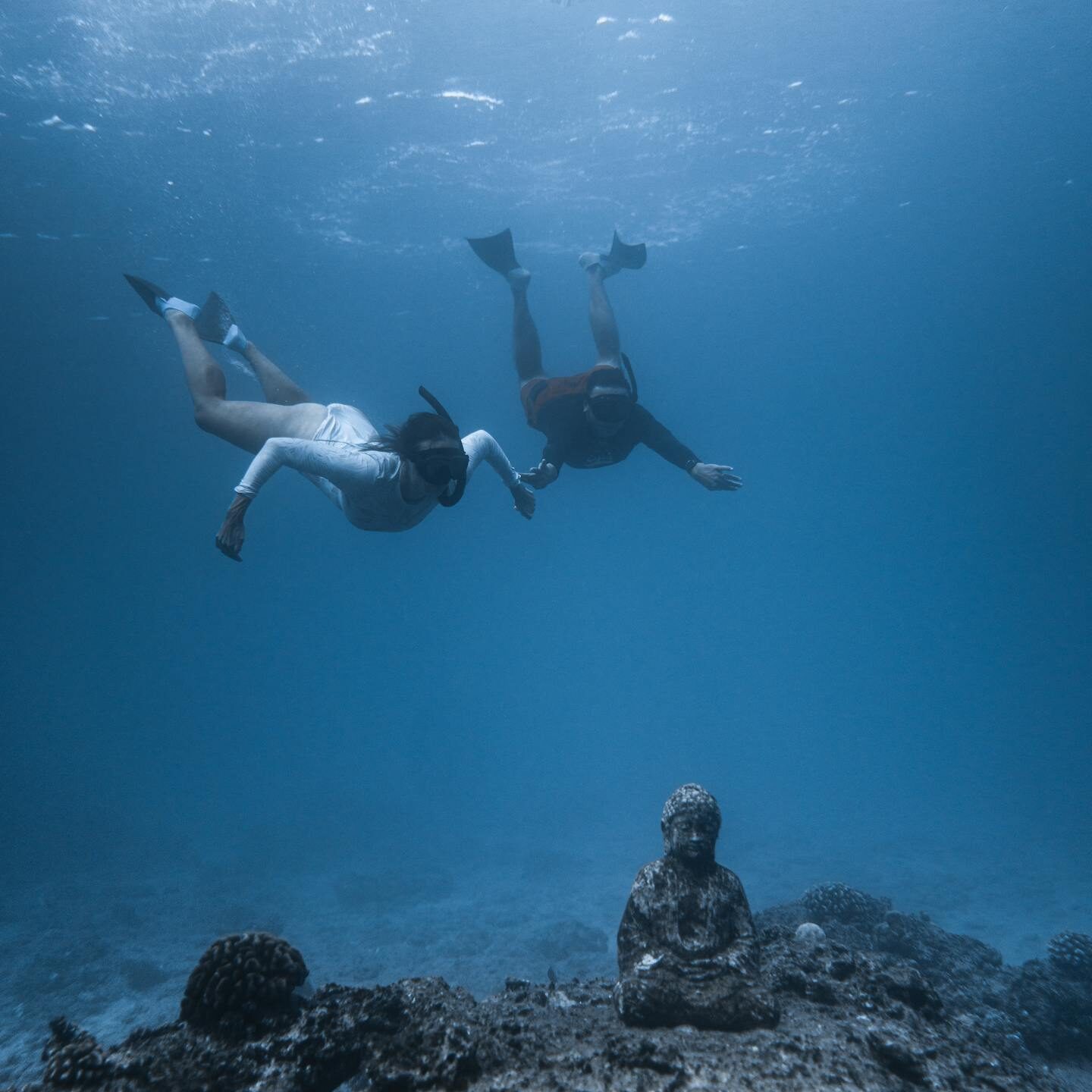 Unrecognizable divers in scuba masks and flippers swimming underwater of blue ocean and observing ancient sunken Buddha statue